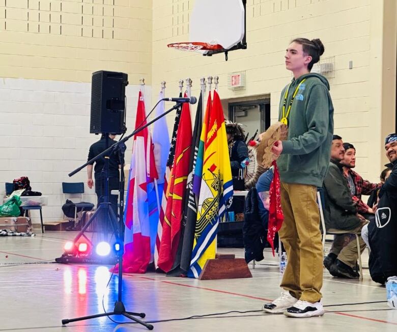 A young man in a green sweater stands in front of a microphone and holds a tiny drum in his hands. 