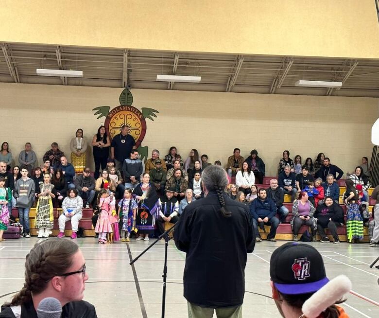 A man wearing a black shirt stands facing away from the camera. In front of him is a microphone and people sitting on bleachers in a gym.