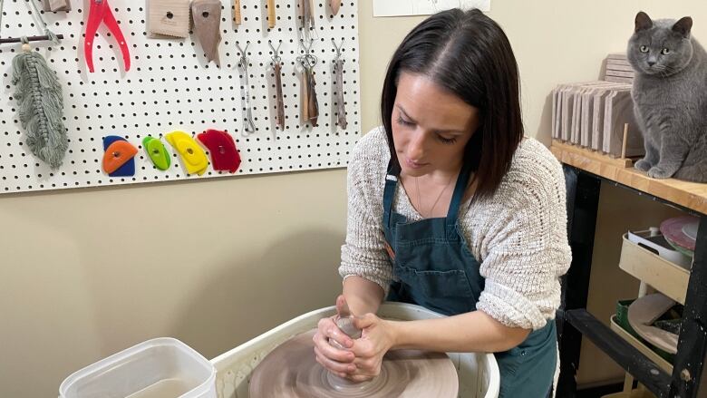 A woman working on clay on a pottery wheel. There is a grey cat standing over her shoulder.