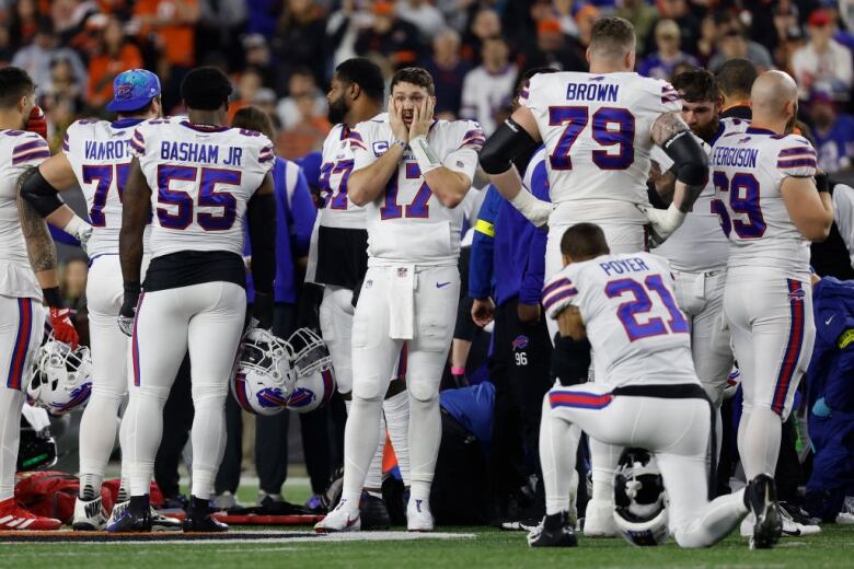 Football players gather on the field amid other personnel. One is holding his face in apparent shock. 