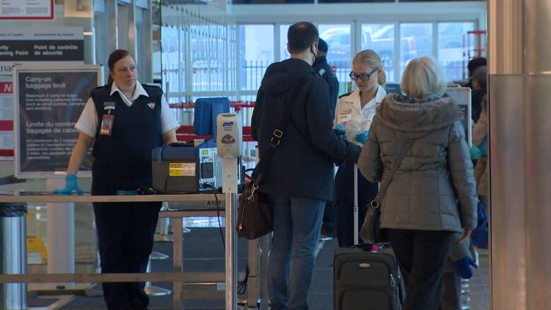 Passengers carrying luggage bags standing in a line. Airport security staff wearing black vests and pants are waiting to check the passengers in. 