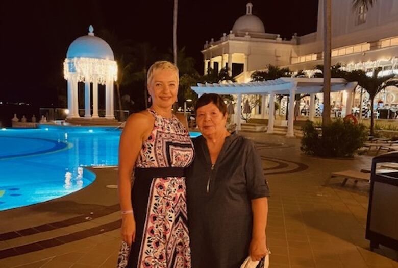 Two women stand next to a swimming pool with palm trees in the background.