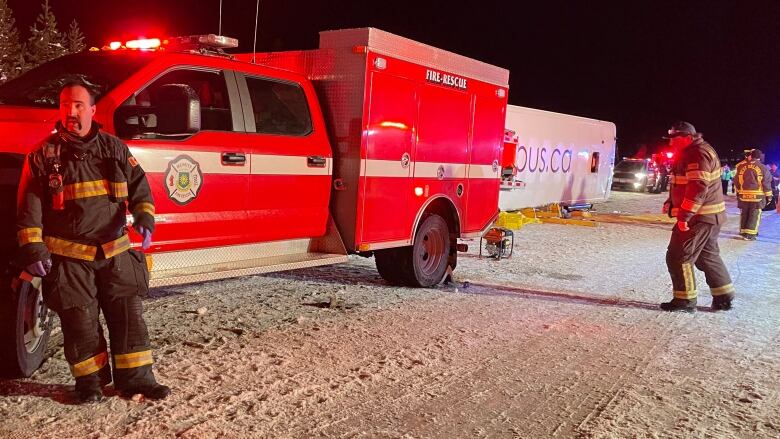 A red rescue truck is pictured in the foreground, behind it a white bus lies toppled over on is side. First responders are on the scene.