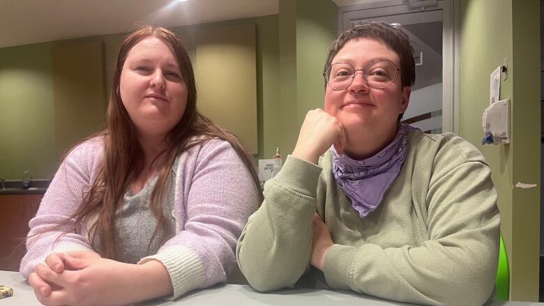 Two women sit beside each other at a boardroom table. One, on the left, has long brown hair and is smiling slightly. The other has short hair, a nose ring, and glasses. She has a big smile and is resting her chin on her hand.