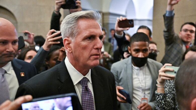 Grey-haired man is surrounded by reporters. 