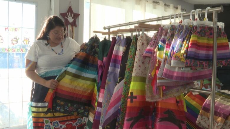 A woman stands behind a row of colourful ribbon skirts. She is also wearing a ribbon skirt and has glasses. 