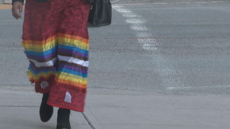 A person wearing a rainbow coloured ribbon skirt walks on the sidewalk. The photo is shot from the waist down. 