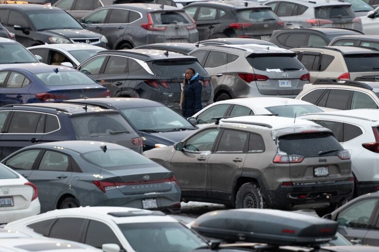 Someone stands in a mall parking lot surrounded by parked, dull-coloured vehicles.