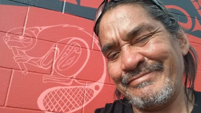 A man takes a selfie in front of a mural featuring Indigenous art.