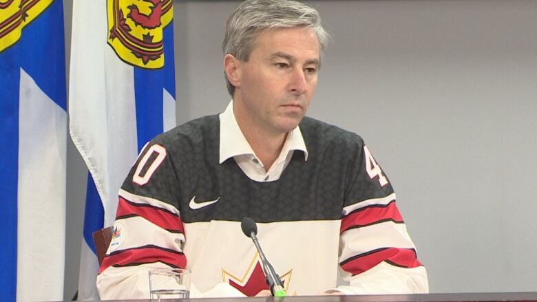 A man with short gray hair and wearing a Team Canada hockey sweater sits as a podium and listens to questions from reporters.