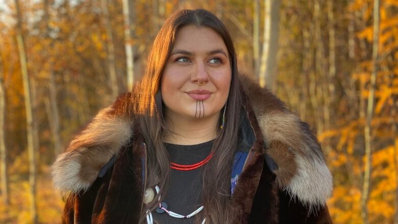 A portrait of a woman with fall foliage in the background. She has traditional tatoo markings on her chin and is wearing traditional necklaces.