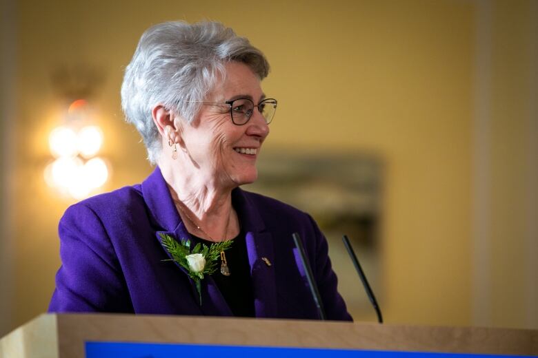 A woman with white hair in a blue blazer stands at a podium and smiles.