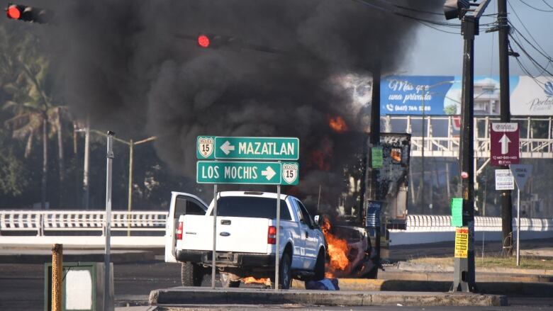 An abandoned white pickup truck sits on a road in front of a burning object which is creating a lot of smoke.