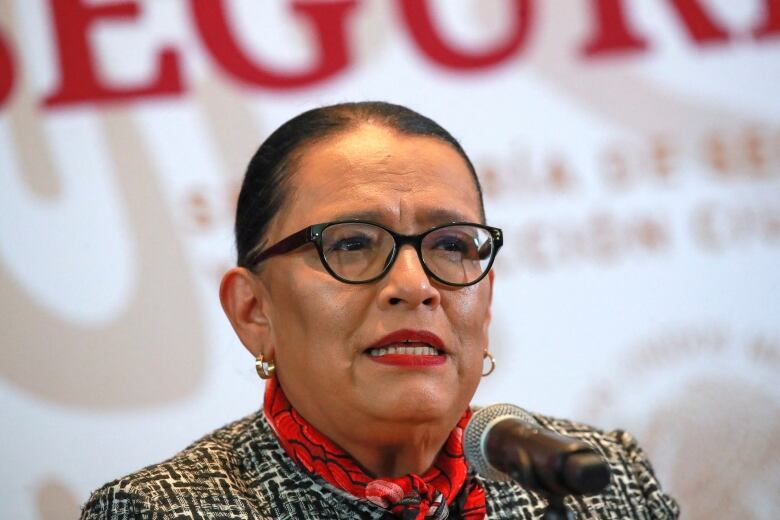 A woman with slicked-back hair and glasses speaks into a microphone.