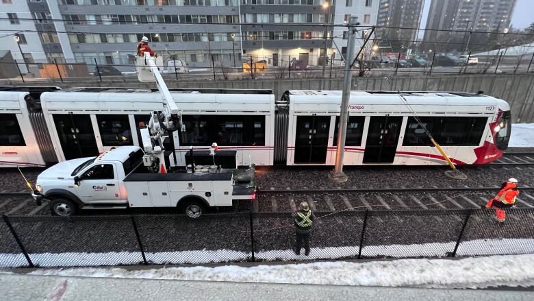 Crews work on an immobile light rail train, seen from above, while towers loom in the distance.