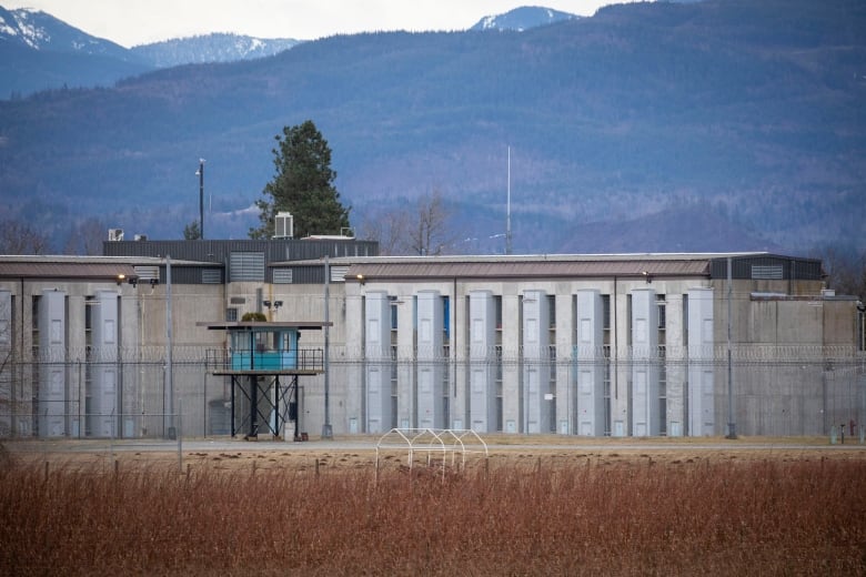 A grey correctional facility building is pictured on an overcast day. Mountains are in the background.