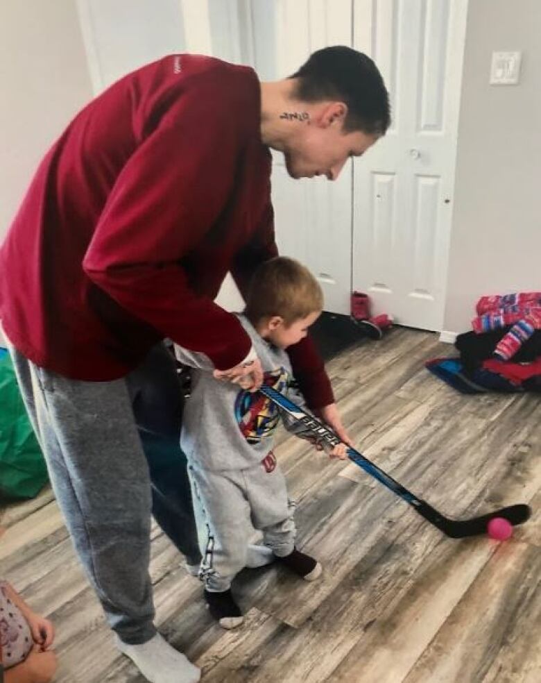 Man stands in living room teaching toddler how to hold hockey stick.