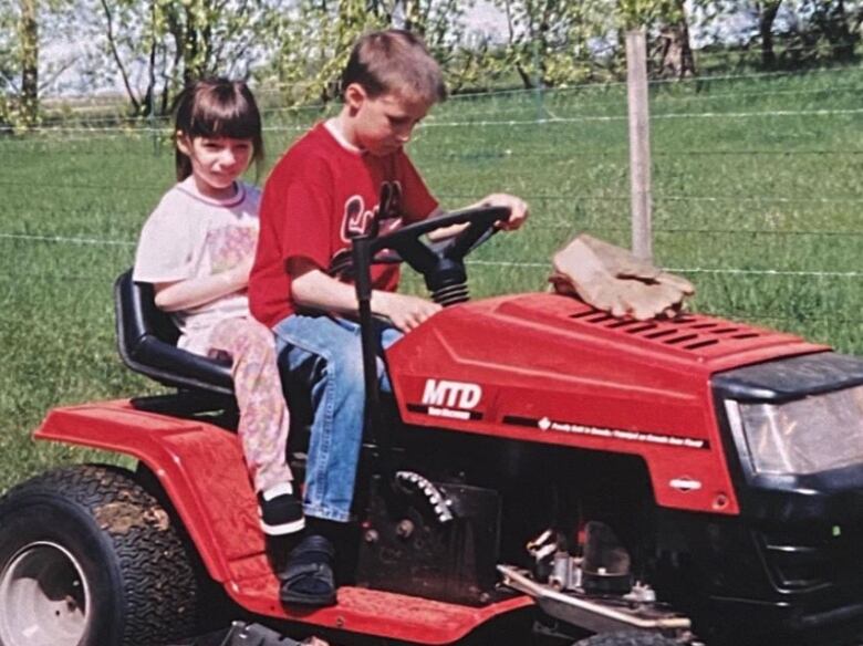 Two young kids driving a riding lawnmower in the summertime.