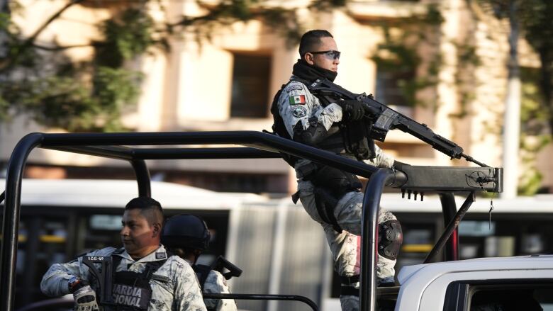 Men holding machine guns wait in a jeep outside a building