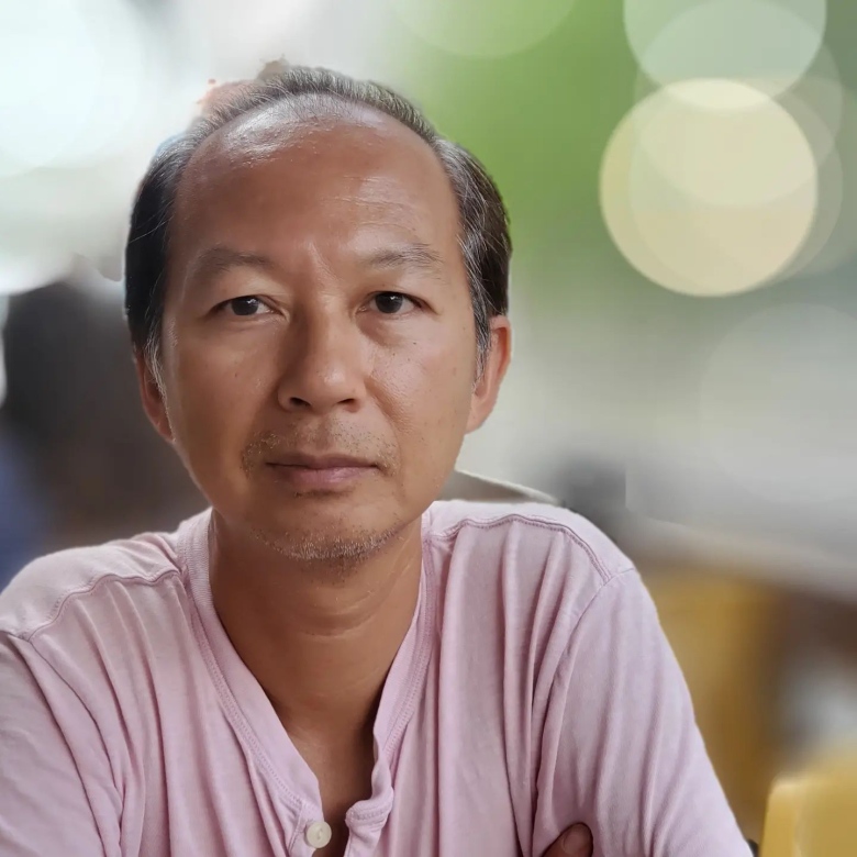 A man wearing a pink shirt looks at the camera with a neutral expression.