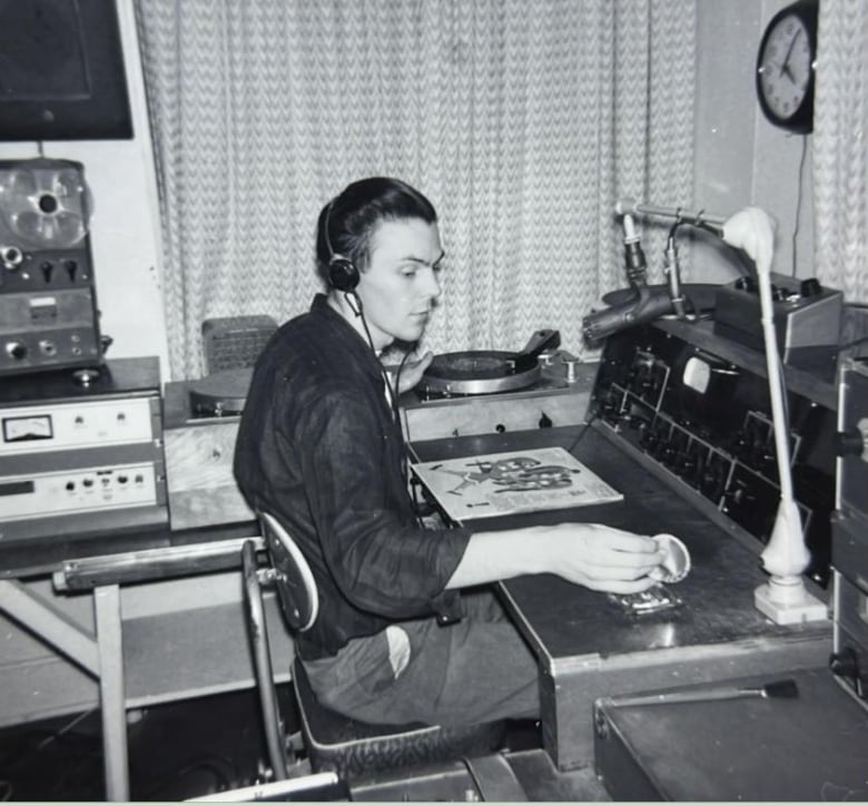 An old photo of a young man seated at a radio console in front of a microphone, preparing to start a turntable to play a record.