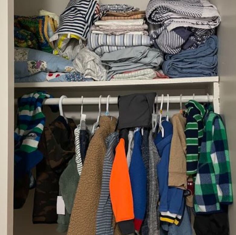 A closet with blankets and sheets on the top shelf and childrens' clothes hanging on the rack.