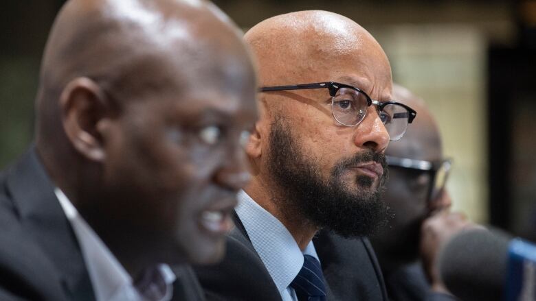 Three men sit next to each other, two men are blurred and the man in the middle is in focus.  