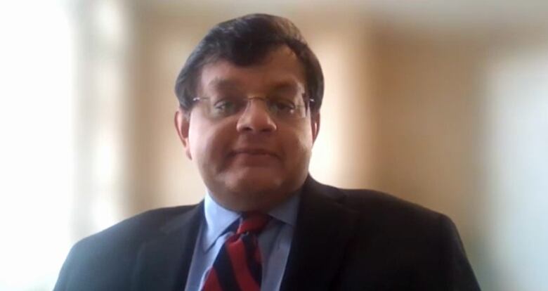 A man with black hair wearing a black suit with a blue shirt and a red and black striped tie and glasses