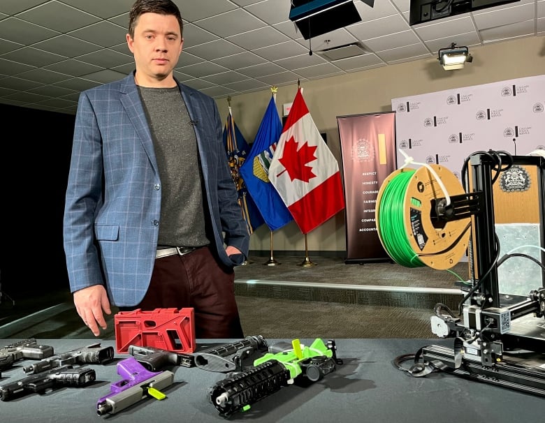 A man in a blue blazer with a grey sweater stands behind a table with guns on it. Some are brightly coloured.