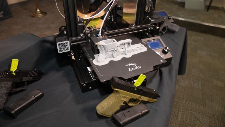 An olive green gun sits on a table with a 3D printer behind it. The 3D printer has a grey firearm on it.