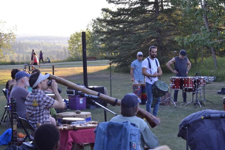 A man speaks to a small crowd while holding a tam tam drum outdoors in a park. 