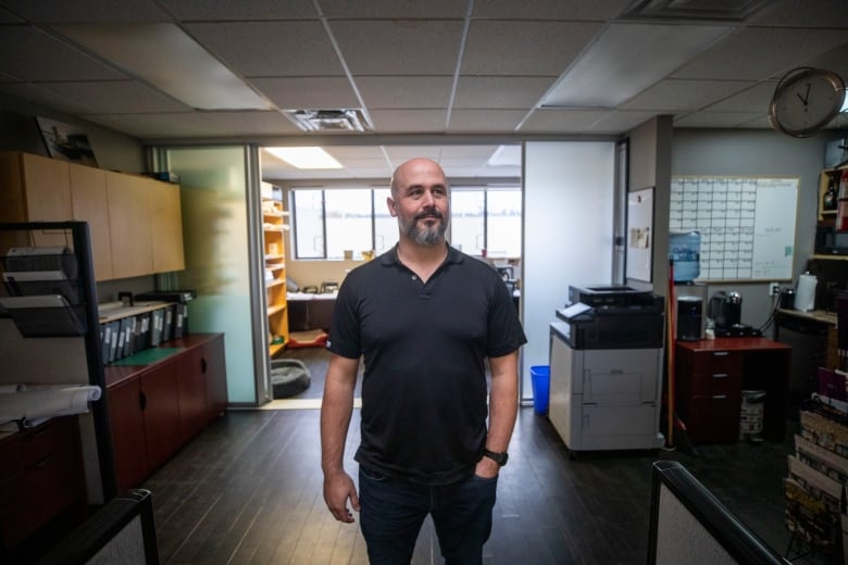 A bearded man wearing an open-necked T-shirt stands in an office with one hand in his pocket.