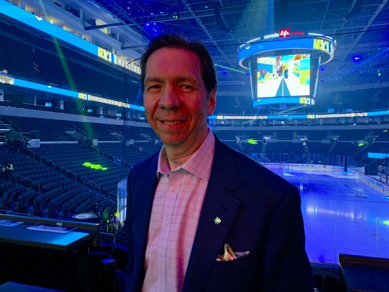 A sportscaster stands in a hockey arena with the ice surface in the background.