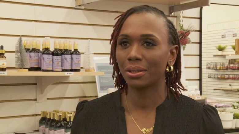 A woman with thin, reddish dreadlocks stands in front of a display of Eleora Beauty products. 