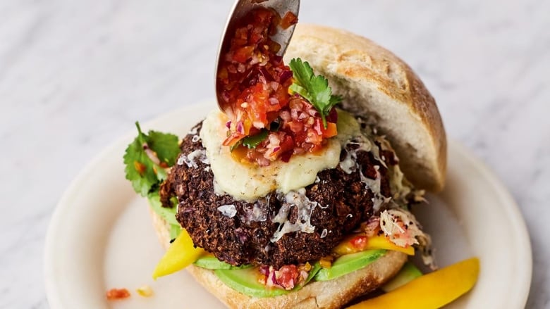 A black bean burger on a bun, sitting on a white plate on top of a white marble surface. A hand reaches into frame to spoon salsa on top of the burger patty.