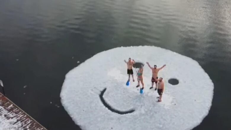 Four people in bathing suits stand on a 10-metre-diameter block of floating ice that they carved into a smiley face.