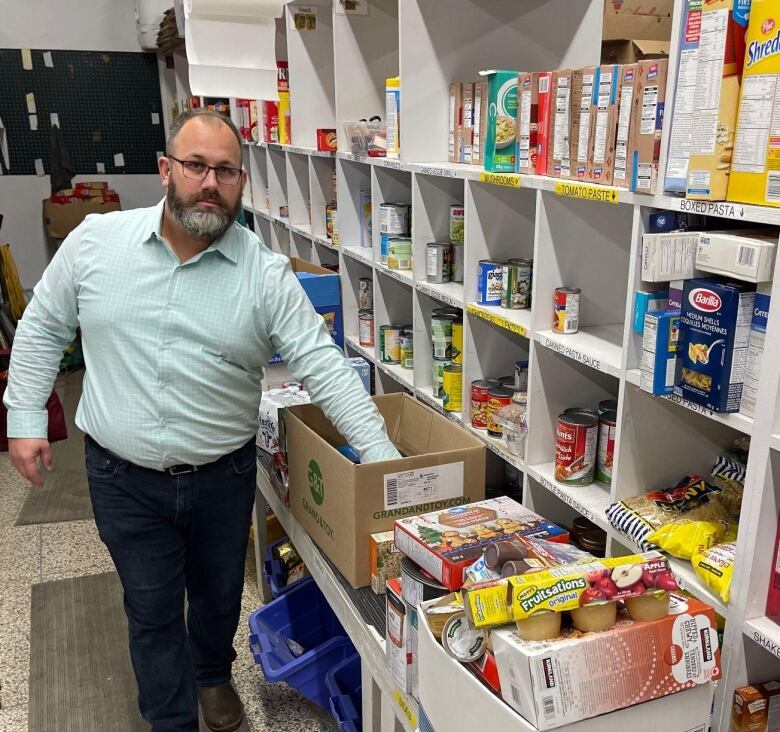 A man bearded man fills a box in a large pantry-style room, stocked with numerous food items. 