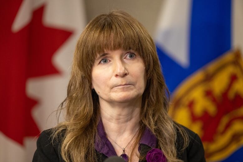 A woman with long hair and bangs is seated and is wearing a purple shirt and a black blazer.