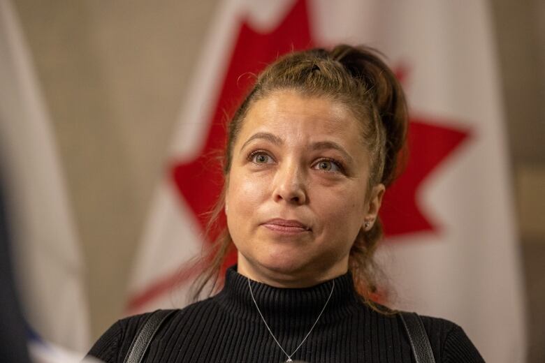 A woman is seated in front of a Canadian flag. Her hair is in a ponytail and she is wearing a black turtleneck.