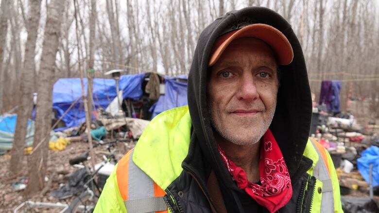 A man in a high-visibility jacket and wearing a handkerchief around his neck stands in front of a shelter made out of tarps that's surrounded by trees.