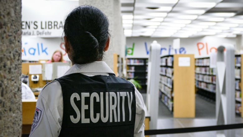 A security guard stands in a library with their back to the camera. The word 