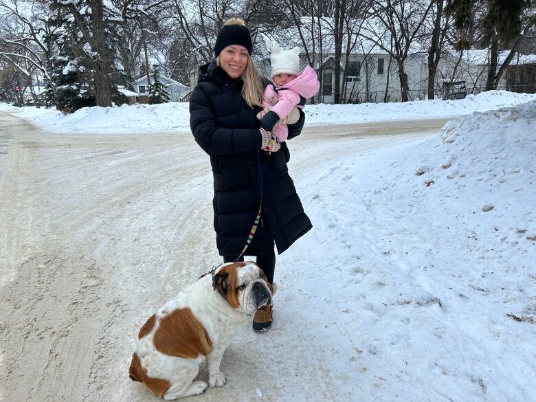 A woman is standing on a street holding a baby and a leash with a bulldog. 