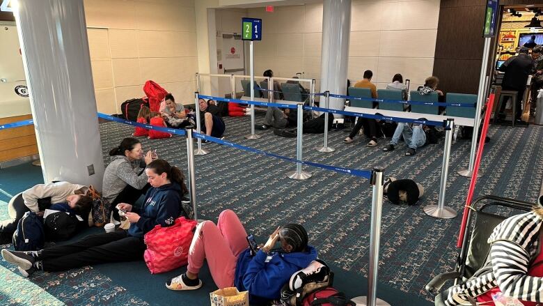 People sit in seats or on the carpeted ground inside an airport.