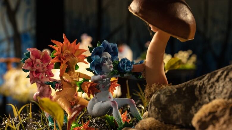 Photo of a detail of a diorama by artist Tammy Salzl. Three polymer clay fairies with flower petals surrounding their heads form a chain and pull on a brown toadstool that is taller than they are. The figures are nude and are in three different colours: one pink, one blue and one yellow.