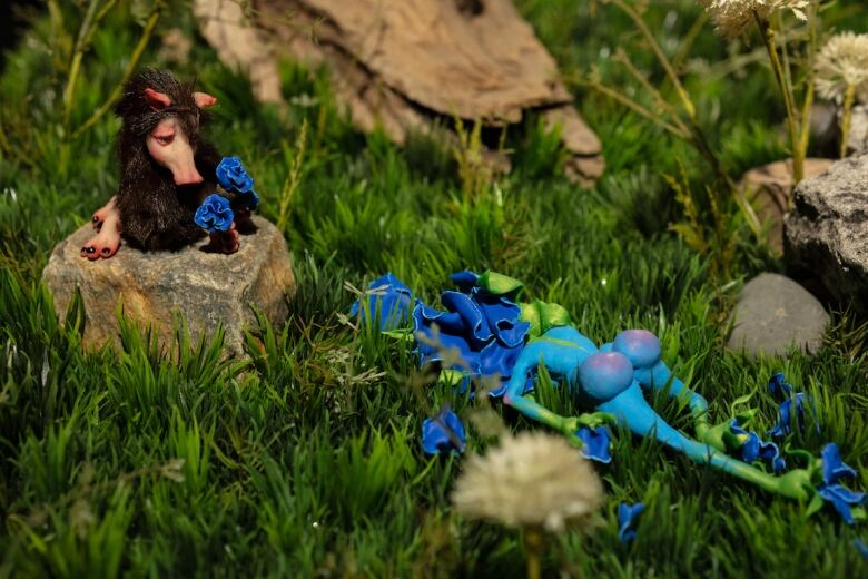 Detail of a diorama by Tammy Salzyl. A tiny sculpture of a nude blue fairy lies dead, face down, in tall green grass. A small and furry figurine of a creature with a pig snout sits on a tree stump next to the fairy, holding a bouquet of blue flowers that have been taken from the fairy's corpse.