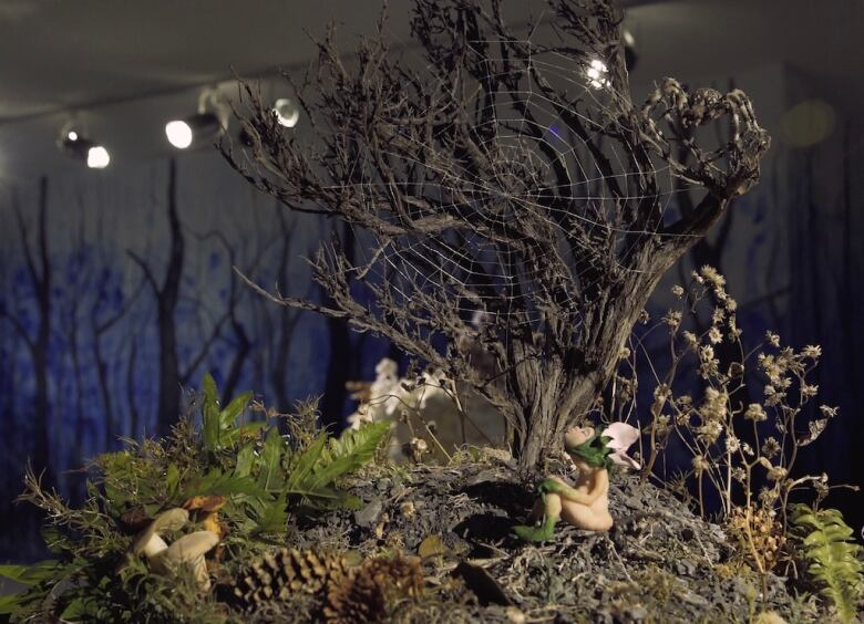 Diorama by Tammy Salzyl. A tiny sculpture of a peach-coloured nude fairy with a pink flower-topped head, sits beneath a gnarled tree with a large cobweb in its branches.