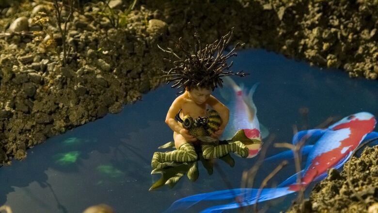 Detail of a diorama by Tammy Salzyl. A tiny figurine of a nude fairy with brambles atop its head, kneels on a video screen that creates the illusion of a fish-filled blue pond. In the fairy's arms is a wasp.