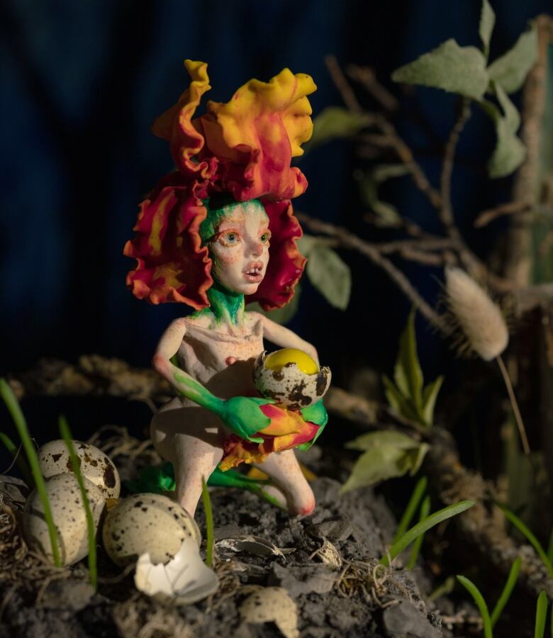 Photo of a miniature clay sculpture of a fairy. It has a fiery flower atop its head and it kneels in a nest of speckled bird's eggs. One broken egg is held in its hands.