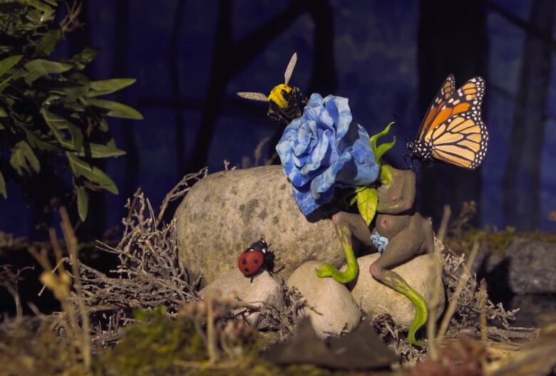 Closeup detail photo of a diorama by Tammy Salzyl. A sculpted clay fairy with grey skin and a blue flower for its head, rests on a pile of stone. Three colourful insects -- a ladybug, butterfly and bee, approach the figure.