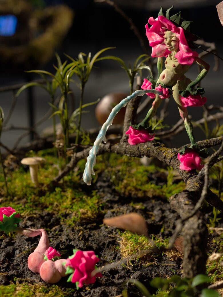 Close-up detail of a diorama by Tammy Salzyl. In a sculpted woodland forest, a nude fairy with pink petals surrounding its face, stands smiling on mossy ground in a power stance. A blue stream of liquid pours from its flowering crotch in a perfect parabolic arch. Pink fairy legs poke from the ground below the arc of fluid, spread eagle.